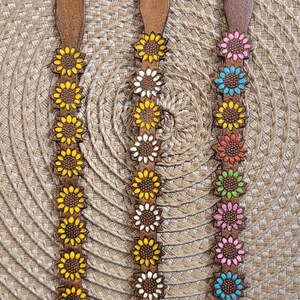 Op de afbeelding: Drie leren armbanden met zonnebloemontwerpen. De armbanden zijn bruin met zilveren sluitingen. De zonnebloemen zijn geel, wit en roze.