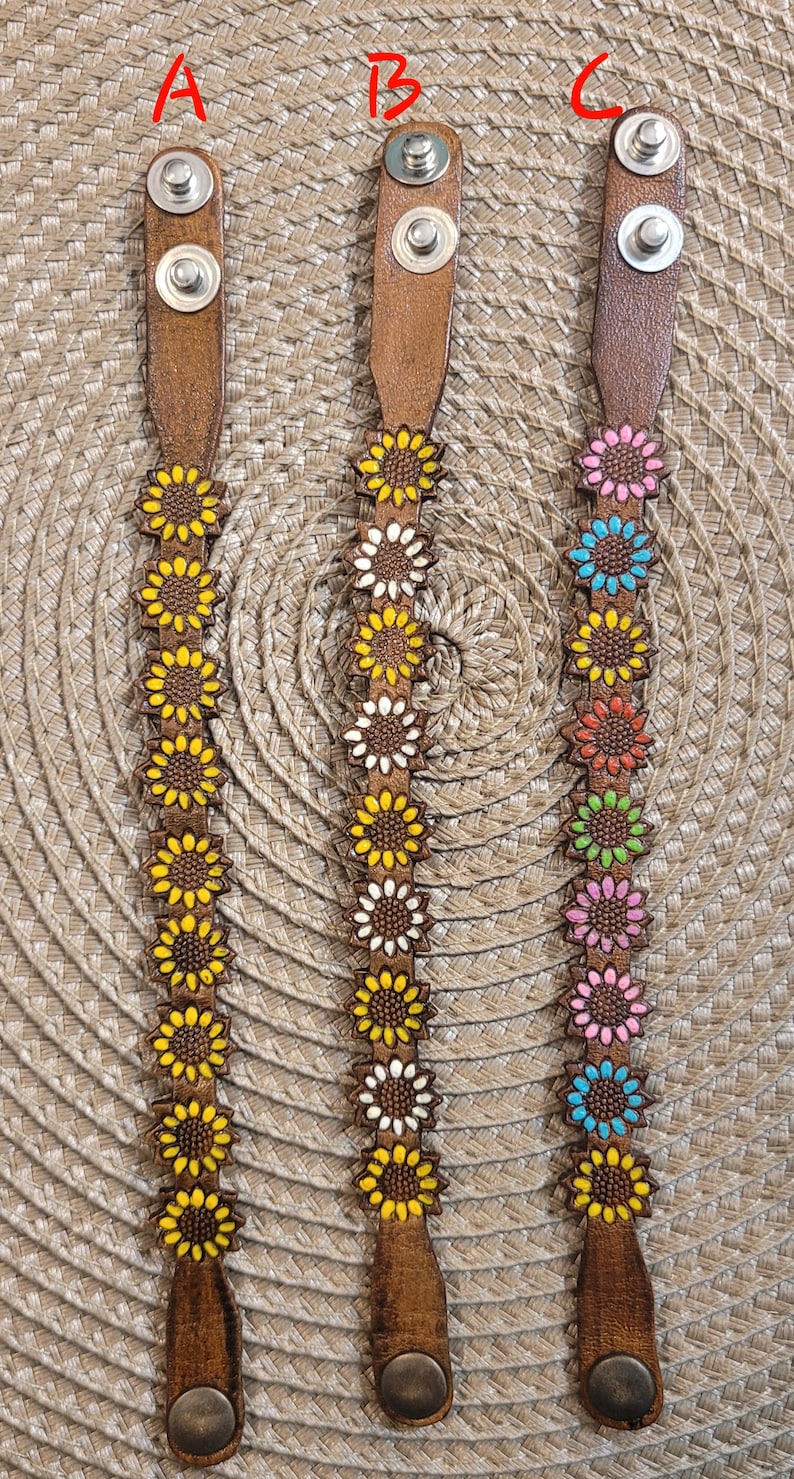 Op de afbeelding: Drie leren armbanden met zonnebloemontwerpen. De armbanden zijn bruin met zilveren sluitingen. De zonnebloemen zijn geel, wit en roze.