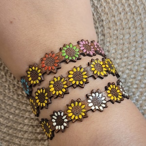 Op de afbeelding: Drie bruine leren armbanden met zonnebloemmotieven. Elke armband heeft een andere kleur zonnebloem: geel, oranje en roze.