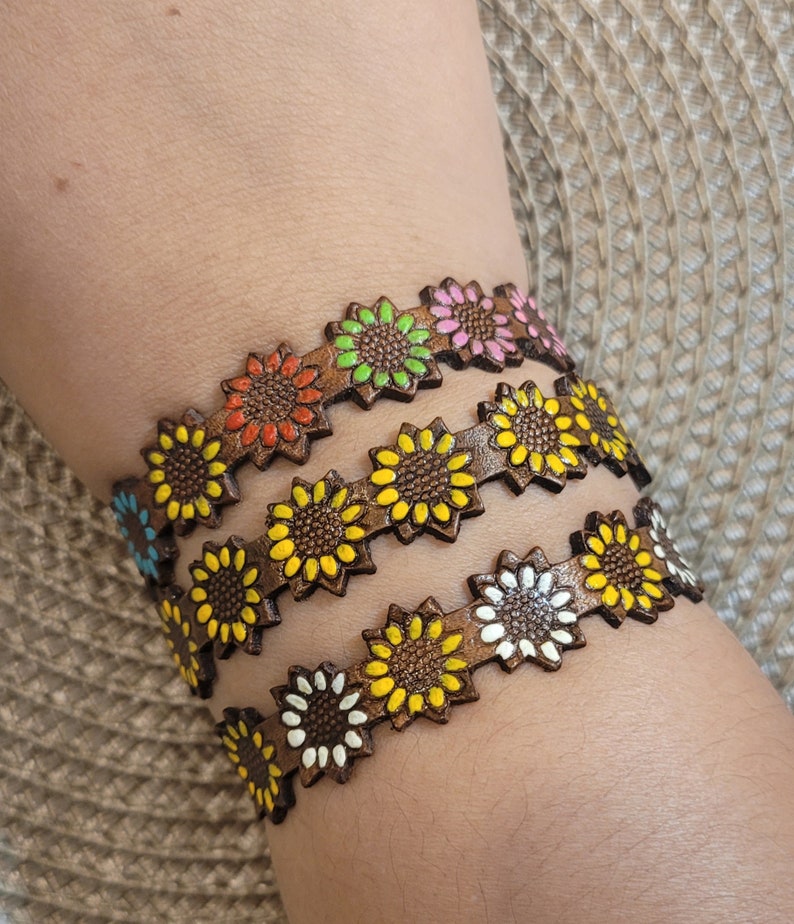 Op de afbeelding: Drie bruine leren armbanden met zonnebloemmotieven. Elke armband heeft een andere kleur zonnebloem: geel, oranje en roze.