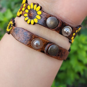 Op de afbeelding: Twee bruine leren armbanden met gele zonnebloemontwerpen en zilveren en bronzen accenten.