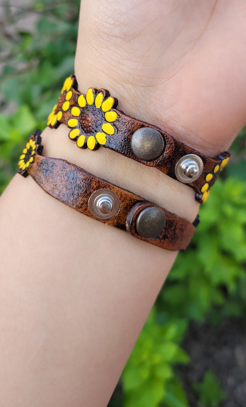 Op de afbeelding: Twee bruine leren armbanden met gele zonnebloemontwerpen en zilveren en bronzen accenten.
