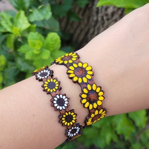 Op de afbeelding: Twee bruine leren armbanden met zonnebloemontwerpen. De ene armband heeft gele zonnebloemen met bruine centra, de andere heeft witte zonnebloemen met bruine centra.