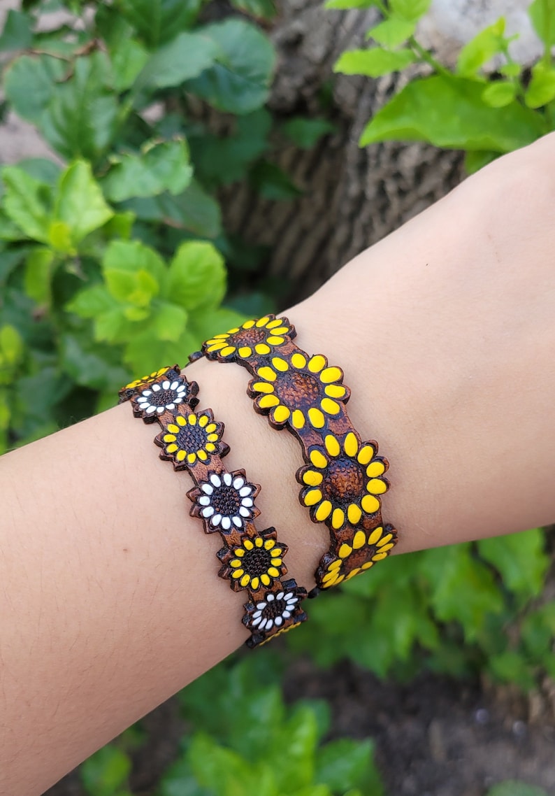 Op de afbeelding: Twee bruine leren armbanden met zonnebloemontwerpen. De ene armband heeft gele zonnebloemen met bruine centra, de andere heeft witte zonnebloemen met bruine centra.