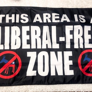 May include: Black flag with white text that reads "This area is a liberal-free zone". The flag has two red circles with a diagonal line through them. Each circle contains a blue donkey with a red star on its side.