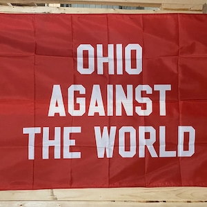 May include: A red flag with the words "OHIO AGAINST THE WORLD" in large, white, block letters. The flag is rectangular and appears to be made of a lightweight fabric. The flag is hanging on a wooden structure.