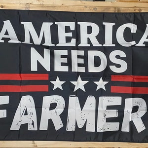 May include: A black flag with white text that reads "America Needs Farmers". There are three white stars and two red stripes on the flag.
