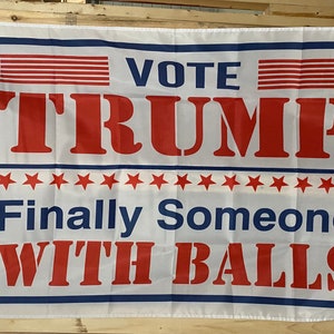 May include: A white flag with blue and red text that reads "Vote Trump Finally Someone With Balls". The text is in a bold, blocky font. There are red stripes and stars on the flag.