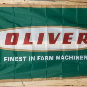 May include: A green and gold flag with the word "OLIVER" in red letters on a white oval. The text "FINEST IN FARM MACHINERY" is printed below the oval.