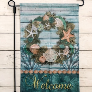 May include: A welcome garden flag with a seashell wreath on a blue and white woodgrain background. The wreath is made of seashells, starfish, and coral. The text "Welcome" is written in gold lettering at the bottom of the flag.