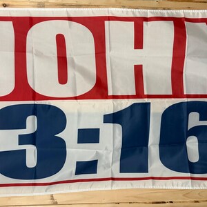 May include: A rectangular flag with the text "JOHN 3:16" in large, bold letters. The word "JOHN" is in red letters on a white background, and "3:16" is in blue letters on a white background. The flag has a red border.