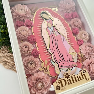 Puede incluir: Una obra de arte enmarcada con un diseño en capas de una figura religiosa en rosa y dorado, rodeada de flores de papel en tonos rosados. El nombre "Daliah" se muestra en una placa de madera en la parte inferior. La obra está encerrada en un marco blanco.