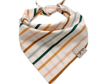 May include: A triangular pet bandana featuring a cream-colored fabric with diagonal stripes in shades of dark green, mustard yellow, and soft pink. A small, square, light-colored leather-like tag is stitched to the corner with the text mjups.