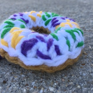 Puede incluir: Una dona de ganchillo con glaseado morado, verde, amarillo y blanco. La dona est&aacute; sobre una superficie de hormig&oacute;n gris.
