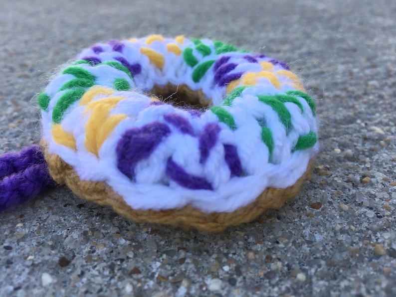 Puede incluir: Una dona de ganchillo con glaseado morado, verde, amarillo y blanco. La dona est&aacute; sobre una superficie de hormig&oacute;n gris.