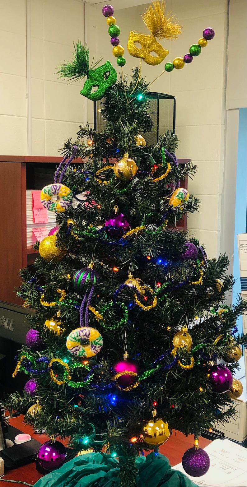 Puede incluir: Un &aacute;rbol de Navidad artificial verde decorado con adornos morados, dorados y verdes, incluyendo m&aacute;scaras de Mardi Gras, cuentas y una estrella dorada en la parte superior.
