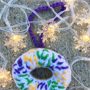 Puede incluir: Un adorno de donut de ganchillo en hilo blanco, morado, verde y amarillo. El adorno est&aacute; decorado con un patr&oacute;n de remolino morado, verde y amarillo. El adorno cuelga de un lazo de hilo morado.