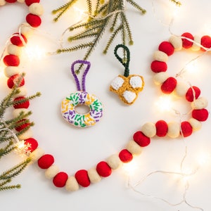 Puede incluir: Dos adornos de crochet, uno con forma de donut con glaseado morado, verde y amarillo y el otro con forma de estrella con hilo blanco y amarillo. Los adornos cuelgan de una cadena de luces blancas y bolas de fieltro rojo y blanco.
