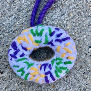 Puede incluir: Una corona de ganchillo en blanco, morado, amarillo y verde. La corona tiene forma de rosquilla y tiene un agujero en el centro. Est&aacute; colgada de un cord&oacute;n morado.