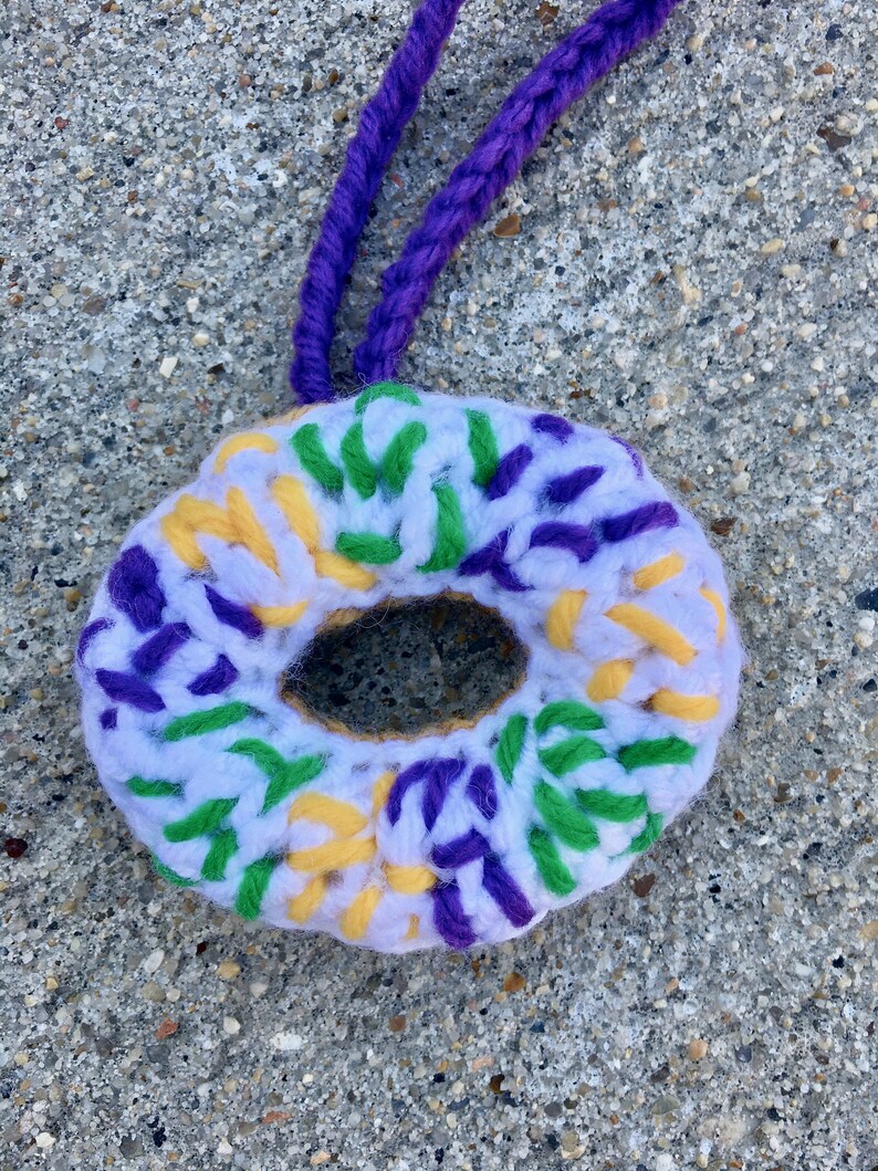 Puede incluir: Una corona de ganchillo en blanco, morado, amarillo y verde. La corona tiene forma de rosquilla y tiene un agujero en el centro. Est&aacute; colgada de un cord&oacute;n morado.