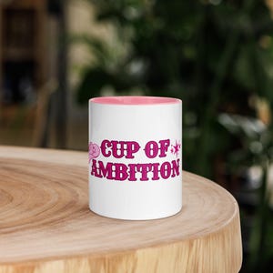 May include: White ceramic mug with a pink interior and the words "CUP OF AMBITION" in pink lettering. The mug is sitting on a wooden surface, with a blurred green background.