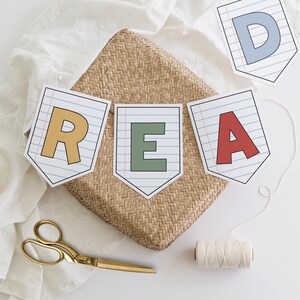Puede incluir: Una guirnalda que deletrea "READ" con letras coloridas en banderines de papel. La guirnalda est&aacute; ensartada con una cuerda blanca y descansa sobre una cesta tejida. Tambi&eacute;n hay unas tijeras doradas y una bobina de hilo.