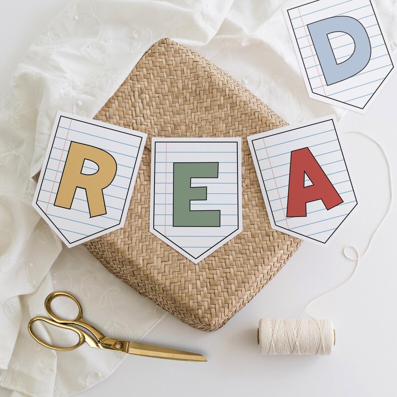 Puede incluir: Una guirnalda que deletrea "READ" con letras coloridas en banderines de papel. La guirnalda est&aacute; ensartada con una cuerda blanca y descansa sobre una cesta tejida. Tambi&eacute;n hay unas tijeras doradas y una bobina de hilo.