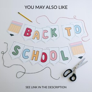 Puede incluir: Una pancarta de "Back to School" con letras coloridas e ilustraciones de l&aacute;pices. La pancarta est&aacute; hecha de papel y est&aacute; ensartada con hilo rojo y blanco. Tambi&eacute;n se incluyen un l&aacute;piz amarillo y unas tijeras.