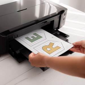 Puede incluir: Una impresora de inyecci&oacute;n de tinta negra imprimiendo una hoja de papel con las letras "E" y "R" en verde y dorado. El papel tiene un patr&oacute;n de cuadr&iacute;cula y est&aacute; siendo sostenido por las manos de una persona.