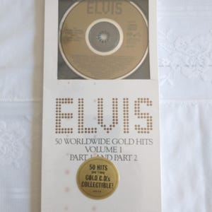 May include: A sealed CD set featuring two gold compact discs. The packaging is white with the name "ELVIS" in a dotted font. Text on the packaging reads "50 Worldwide Gold Hits Volume 1 Part 1 and Part 2". A gold sticker reads "50 Hits on Two Gold C.D.'s Collectible!"