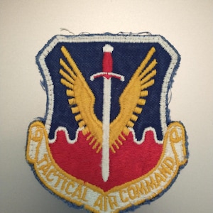 Può includere: Toppa ricamata con bordo blu e disegno a scudo. La toppa presenta una spada rossa, ali gialle e la scritta "TACTICAL AIR COMMAND" in giallo. Lo sfondo è blu e rosso.