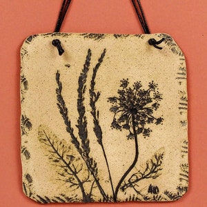 May include: A beige ceramic tile with a black and white pressed flower design. The tile has a rustic look and is perfect for adding a touch of nature to your home decor.