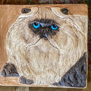 May include: A ceramic tile with a relief sculpture of a white Persian cat with blue eyes. The cat is looking directly at the viewer with a serious expression. The tile is brown and has a textured surface.