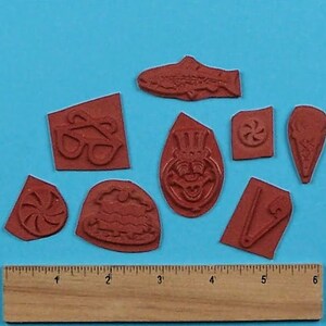 May include: A set of nine red rubber stamps with various designs, including a fish, a pair of glasses, a cake, a candy cane, a pin, a face with a top hat, and a swirl design. The stamps are arranged on a blue surface with a wooden ruler.