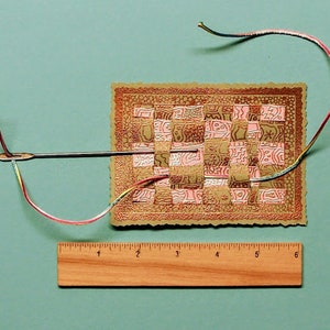 May include: A close-up of a woven tapestry with a pink and brown pattern. A needle and thread are threaded through the tapestry, and a wooden ruler is placed below it.