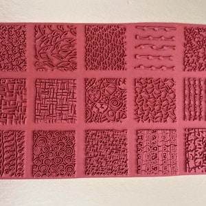 May include: A pink silicone texture sheet with fifteen unique square patterns. The patterns include floral, leaf, and geometric designs, as well as wavy lines and textured grids. Ideal for crafting and adding detail to various projects.