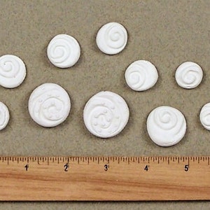May include: Nine white ceramic ornaments with a swirl design. The ornaments are arranged in a row on a brown surface.