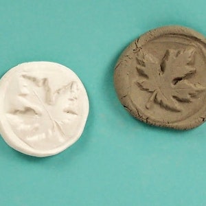 Puede incluir: Dos impresiones redondas de arcilla de una hoja de arce. Una impresión es blanca y la otra es marrón.