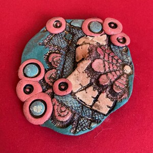 May include: A handcrafted, abstract brooch with a teal base, adorned with coral-colored circles and textured details. The design incorporates beige and pink elements, creating a unique, artistic accessory. The brooch is set against a red background.