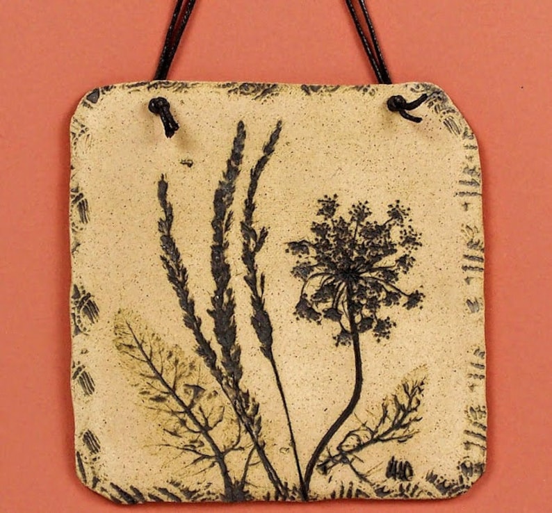 May include: A beige ceramic tile with a black and white pressed flower design. The tile has a rustic look and is perfect for adding a touch of nature to your home decor.