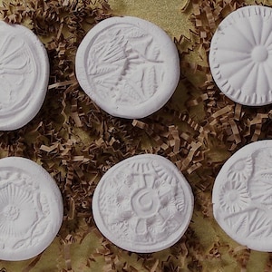 以下が含まれることがあります： 異なる浮き彫りのデザインが施された白い粘土の円盤が6枚。デザインには、花、シダ、太陽、幾何学模様が含まれています。