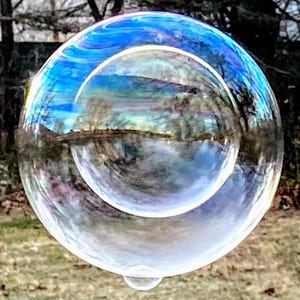Könnte beinhalten: Eine große, schillernde Seifenblase schwebt in der Luft und reflektiert die Umgebung. Die Oberfläche der Blase schimmert in Blau-, Grün- und Weißtönen, mit einer kleineren Blase im Inneren. Der Hintergrund zeigt Bäume und eine grasbewachsene Fläche.