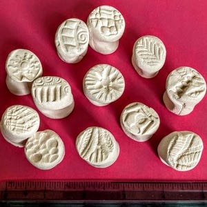 May include: A collection of eleven small, off-white ceramic stamps with various embossed designs, including geometric patterns and stylized images. The stamps are arranged on a red surface, with a ruler at the bottom.