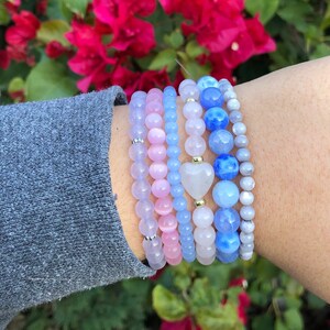 Puede incluir: Un conjunto de seis pulseras con cuentas rosas, azules y grises. Las pulseras est&aacute;n hechas de diferentes materiales, incluyendo vidrio, metal y piedra. Las pulseras est&aacute;n apiladas en la mu&ntilde;eca de una persona.