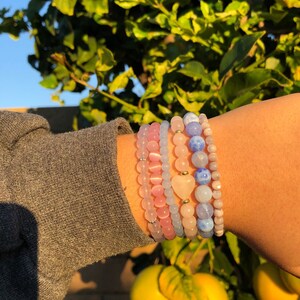 Puede incluir: Un conjunto de cinco pulseras con cuentas rosas, azules y blancas. Las pulseras est&aacute;n superpuestas en la mu&ntilde;eca de una persona y tienen un peque&ntilde;o dije en forma de coraz&oacute;n.
