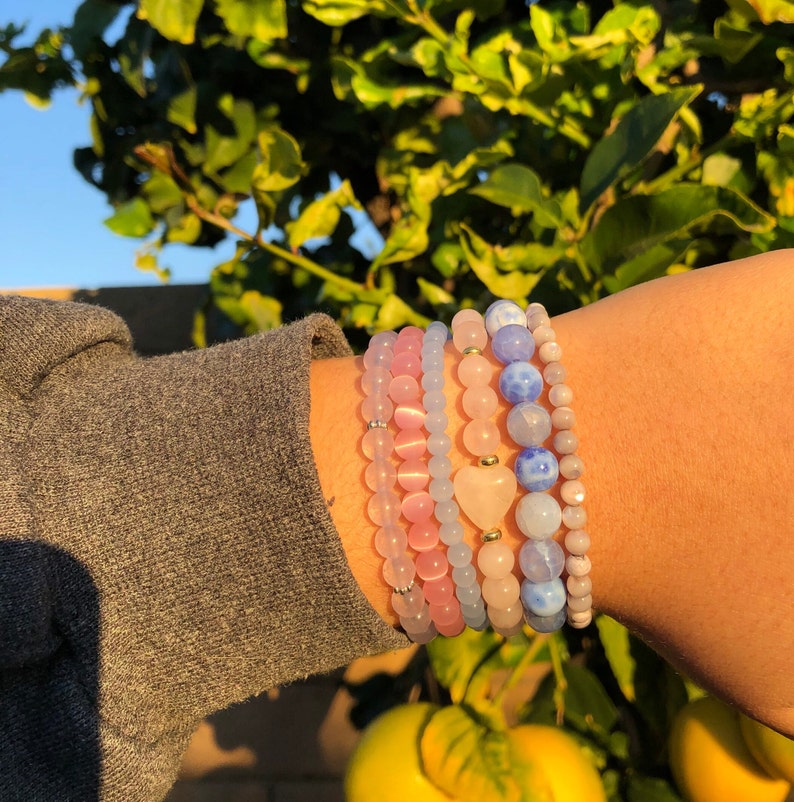 Puede incluir: Un conjunto de cinco pulseras con cuentas rosas, azules y blancas. Las pulseras est&aacute;n superpuestas en la mu&ntilde;eca de una persona y tienen un peque&ntilde;o dije en forma de coraz&oacute;n.