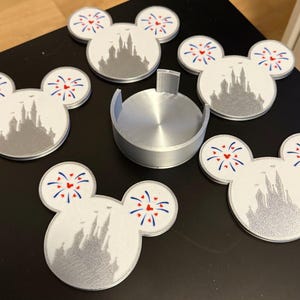 Könnte beinhalten: Set mit sechs weißen und silbernen Mickey-Mouse-Untersetzern mit einer Schloss-Silhouette. Jeder Untersetzer hat rote, weiße und blaue Feuerwerksakzente. Ein silberner Untersetzerhalter ist ebenfalls enthalten.