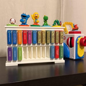 Könnte beinhalten: Ein weißes Regal mit Miniaturfiguren von Sesame Street-Charakteren und kleinen Büchern. Die Bücher tragen Titel wie "My First Pet" und "Tootle". Rechts befindet sich eine bunte Spielzeugkamera.