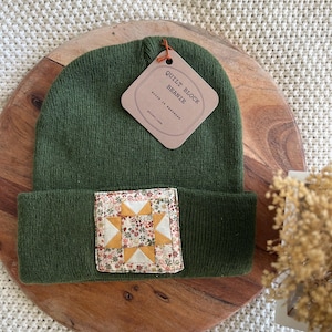 May include: A dark olive green knit beanie featuring a square fabric patch with a yellow and cream star quilt block pattern on a floral background. A brown paper tag attached to the beanie reads "QUILT BLOCK BEANIE" and "BLOCK IS HANDMADE".