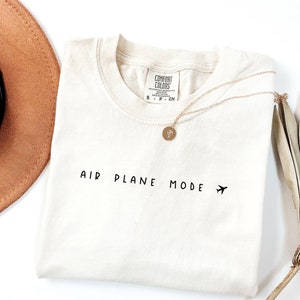 Peut inclure: Un t-shirt blanc avec le texte "AIR PLANE MODE" et une petite ic&ocirc;ne d'avion imprim&eacute;e sur le devant. Le t-shirt est pli&eacute; et pos&eacute; &agrave; plat sur une surface blanche.